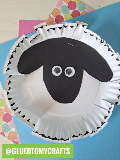 A sheep craft made from a white paper plate with cut fringes around the edge, black paper for the face and ears, and two googly eyes. Colorful polka-dotted paper is in the background.