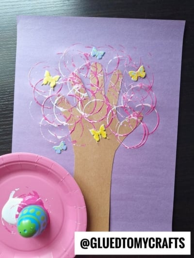 A purple paper features a brown cutout of a hand and arm, decorated with pink and white paint circles and butterfly stickers, resembling a spring tree; a pink paint-filled plate and a colorful roller are nearby.