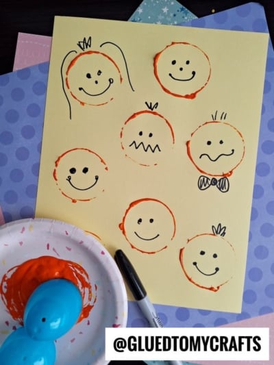 A yellow paper with orange stamped circles, each forming a face with different expressions drawn in black marker. A paint-covered plate, blue plastic eggs, and papers are nearby. Text: @GLUEDTOMYCRAFTS.