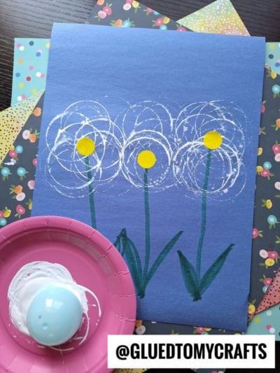 A blue paper features three dandelion flowers painted with white circles and yellow centers using a round stamp. Green stems and leaves are drawn below. A pink plate with white paint and a blue stamp sits beside the artwork. Patterned papers are underneath.