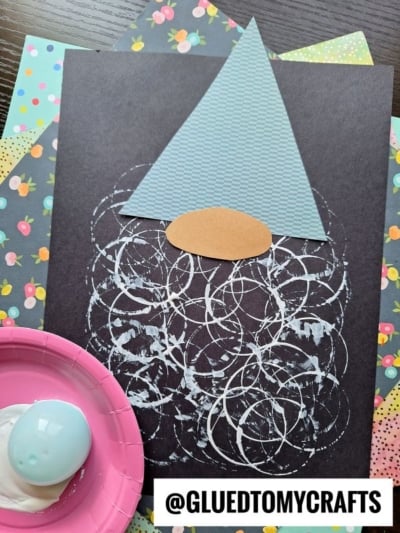 A gnome craft made from patterned paper: a blue triangle hat, a brown oval nose, and a beard of white paint circles on black paper. Colorful floral craft papers and a pink plate with a painted egg are nearby.