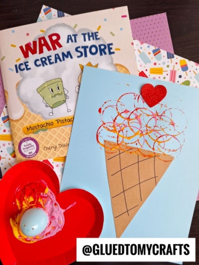 A handmade ice cream cone card with a red heart sits on blue paper, next to the book War at the Ice Cream Store and a heart-shaped tray for Plastic Egg Stamp Crafts. Colorful patterned papers are underneath.