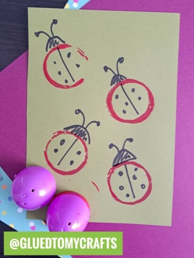 A sheet of green paper features four ladybug drawings made with black and red marker. Two purple plastic eggs are in the corner, and the handle @GLUEDTOMYCRAFTS is written on a green label at the bottom.