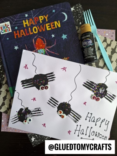 A handmade Halloween card with black spider crafts—perfect for your next INSECT CRAFT IDEAS project—lies next to a Happy Halloween book, black paint, and craft supplies. The card says Happy Halloween and features stars.