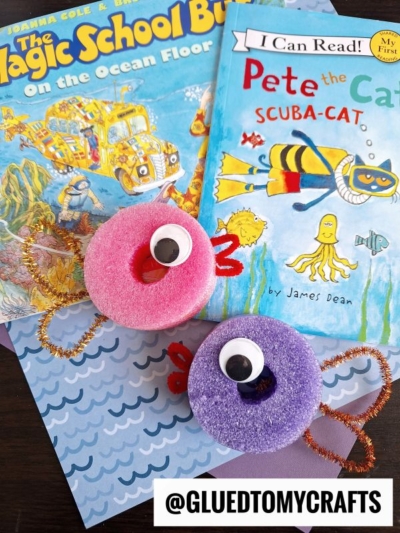 Two children’s books, The Magic School Bus on the Ocean Floor and Pete the Cat: Scuba-Cat, are shown with two colorful DIY sea creature crafts made from sponges, googly eyes, and pipe cleaners.