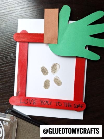 A handmade card with a red craft stick frame, a green paper hand cutout, and brown paper, featuring fingerprint art in the center and the words LOVE YOU TO THE CORE written on the frame. @GLUEDTOMYCRAFTS is at the bottom.