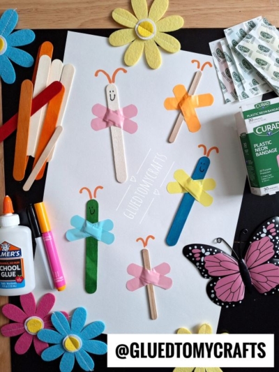 Colorful paper butterflies made with craft sticks, paper flowers, bandages, glue, and markers create fun INSECT CRAFT IDEAS. A butterfly decoration sits on the table, along with the Instagram handle @GLUEDTOMYCRAFTS at the bottom.