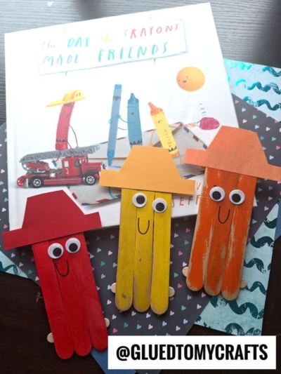 Three colorful popsicle stick crafts with hats and googly eyes are displayed in front of the book The Day the Crayons Made Friends—perfect for back to school story time. Decorative papers are in the background, with the @GLUEDTOMYCRAFTS watermark at the bottom.