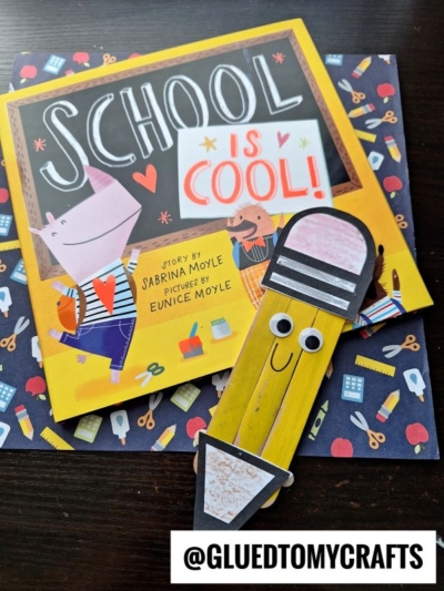 A children’s book titled “School Is Cool!” rests on colorful school-themed paper, perfect for back to school story time, next to a yellow, smiling pencil craft with googly eyes. The handle @GLUEDTOMYCRAFTS is labeled at the bottom.