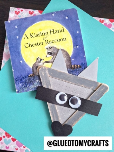 A children’s book titled A Kissing Hand for Chester Raccoon lies next to a handmade raccoon craft made from craft sticks, googly eyes, and paper shapes—perfect for back to school story time on a heart-patterned paper background.