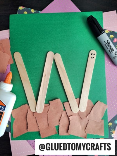 Craft project using popsicle sticks arranged in the shape of a W on green paper with brown paper scraps and glue; a Sharpie marker and floral paper are nearby. @GLUEDTOMYCRAFTS is labeled at the bottom.