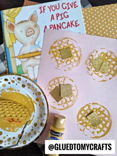 A children’s book titled If You Give a Pig a Pancake, a pink paper with gold painted pancake prints created using a potato masher stamped craft, glittery squares, a paintbrush, and gold paint are arranged on a table. @GLUEDTOMYCRAFTS text is at the bottom.