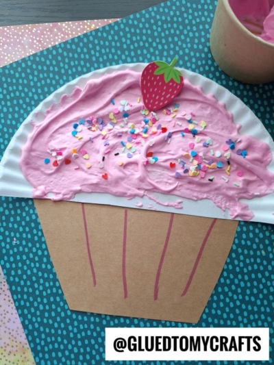 A paper craft cupcake made with a brown paper base and a paper plate top decorated with pink puffy paint, colorful sprinkles, and a paper strawberry on a blue polka dot background—a fun idea for puffy paint crafts.