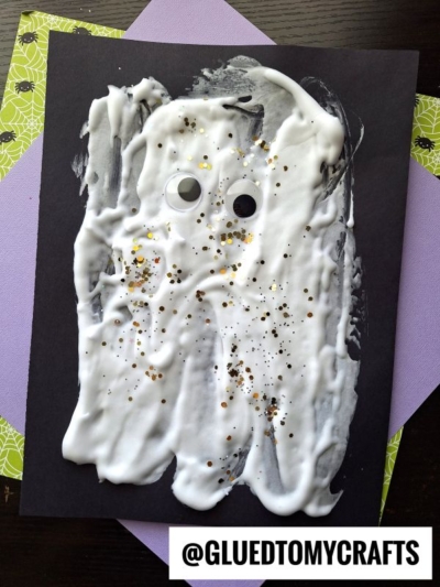A ghost made from white glue and glitter, inspired by puffy paint crafts, with two googly eyes on black paper, resting atop purple and green papers. @GLUEDTOMYCRAFTS is printed at the bottom.