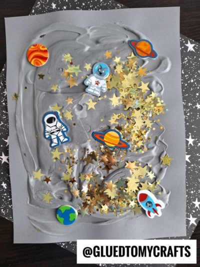 A gray paper with swirled patterns features space-themed stickers of astronauts, planets, and a rocket, plus gold star confetti. Inspired by puffy paint crafts, the handle @GLUETOMYCRAFTS is at the bottom.