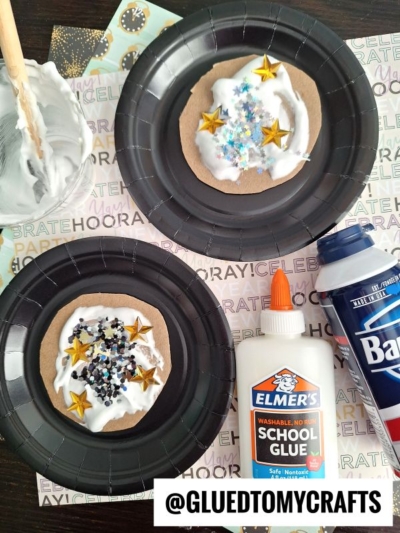 Two black plates hold brown paper circles topped with DIY puffy paint crafts made from white glue, shaving cream, and colorful star and snowflake confetti. Elmer’s glue, shaving cream, and crafting tools are nearby. The text @GLUEDTOMYCRAFTS appears at the bottom.