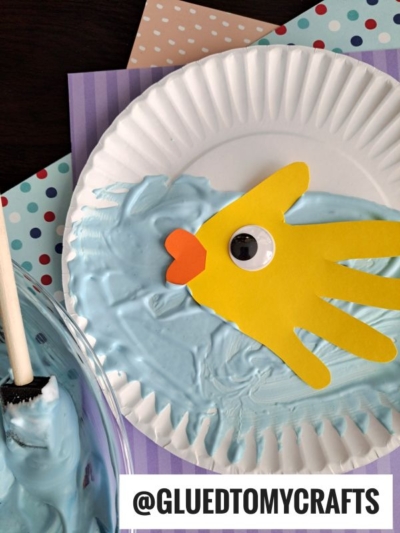 A paper plate features light blue paint resembling water, with a yellow paper hand cutout shaped like a fish, orange paper lips, and a large googly eye; craft materials are visible nearby.