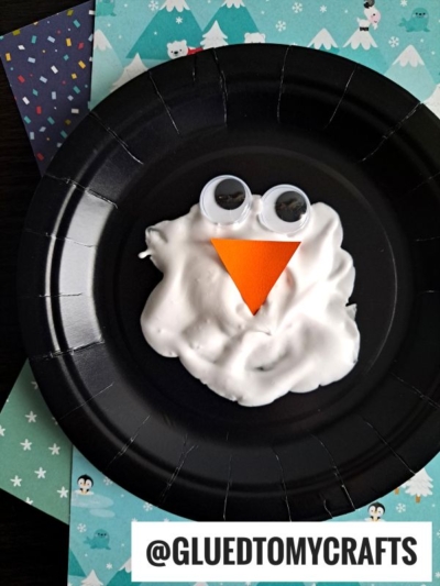 A black paper plate with white puffy paint shaped like a snowman’s face, two large googly eyes, and an orange paper triangle for a nose—perfect for winter puffy paint crafts. The plate sits on patterned papers. @GLUEDTOMYCRAFTS text appears at the bottom.