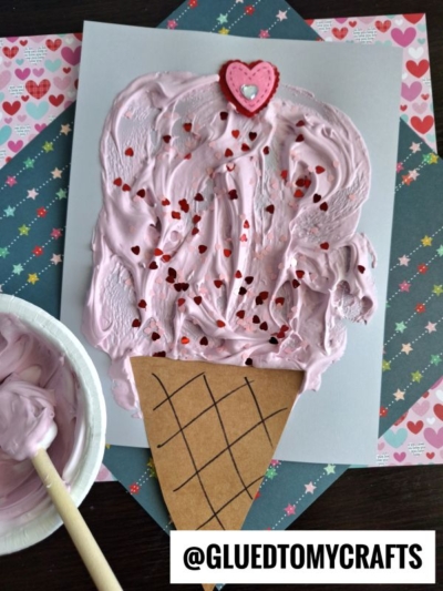 A handmade ice cream craft features a pink, textured scoop with heart-shaped sprinkles, topped with a heart decoration, on a brown paper cone. Puffy paint crafts and pastel heart-patterned paper are in the background.