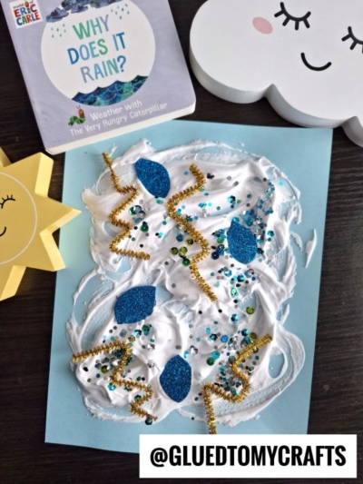 A fun puffy paint crafts project with blue paper decorated in white puffy paint, blue glitter, sequins, and gold pipe cleaners, surrounded by a sun, a cloud, and the children’s book “Why Does It Rain?”.