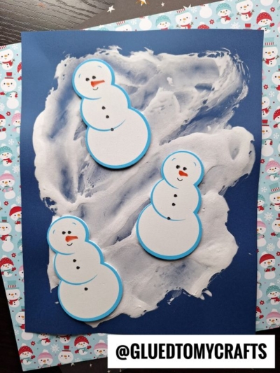 Three paper snowmen with blue edges are glued onto dark blue paper, accented by puffy paint crafts for snowy effects. The background features a repeating snowman pattern. The text @GLUEDTOMYCRAFTS is at the bottom.