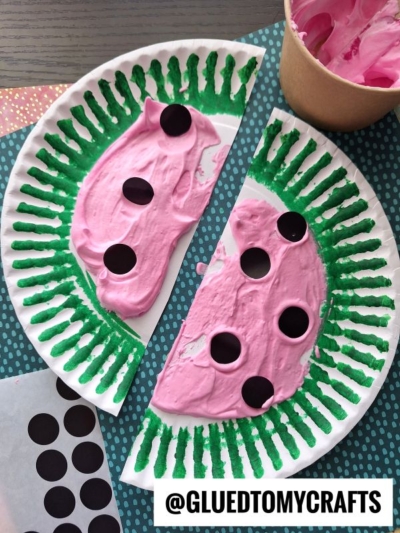 A paper plate painted to look like a watermelon using puffy paint crafts—green for the rind and pink with black paper dots for seeds—cut in half and displayed on a table. The handle @GLUEDTOMYCRAFTS appears at the bottom.