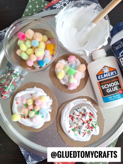 A craft setup with cardboard circles topped with white glue, colorful pom-poms, holiday sprinkles, and touches of puffy paint crafts. Nearby are a bowl of pom-poms, fake snow, Elmers glue, and shaving cream. @GLUEDTOMYCRAFTS is shown at the bottom.