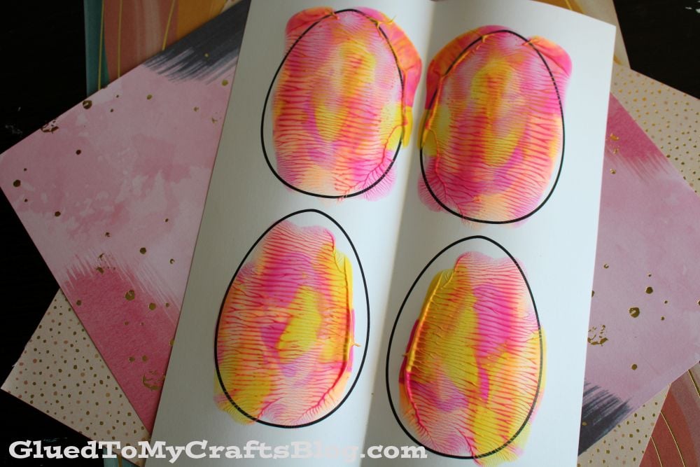 A white paper displays four egg shapes with colorful, swirled paint patterns in pink, yellow, and orange. The paper is placed on top of pink and patterned sheets. GluedToMyCraftsBlog.com is visible at the bottom.