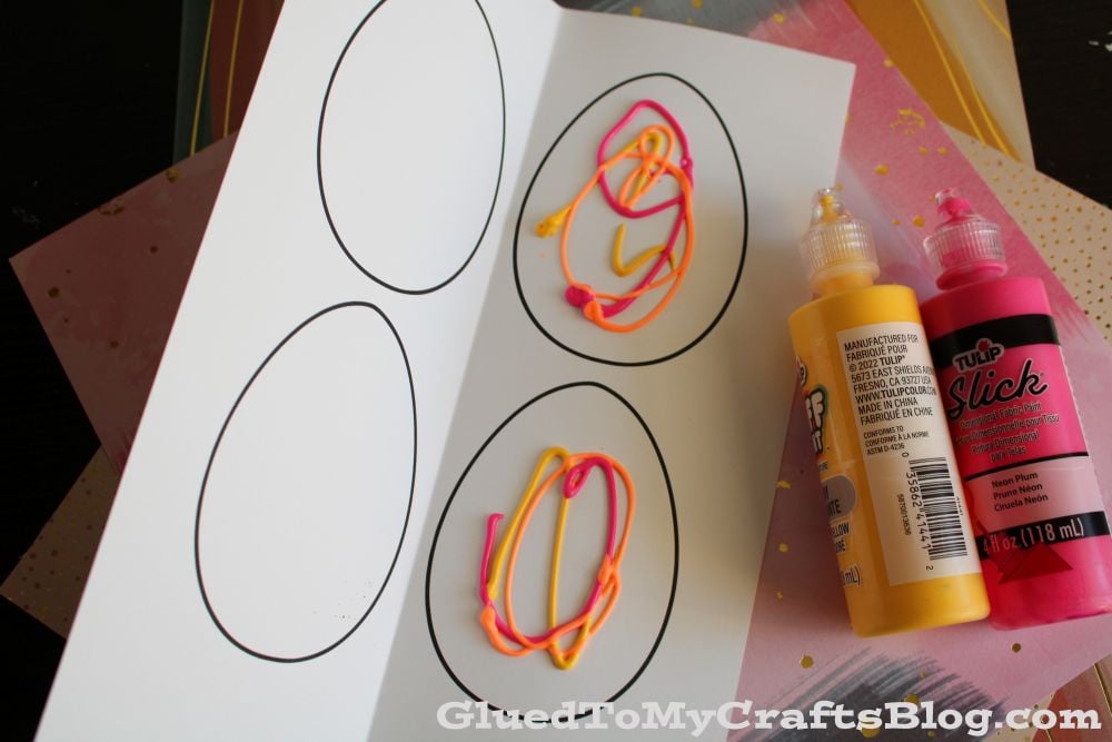 An open card shows three outlined egg shapes, two decorated with yellow and pink puff paint squiggles. Two bottles of puff paint, one yellow and one pink, are next to the card along with sheets of colored paper.
