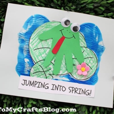 A handmade craft of a green frog with googly eyes and a red tongue, made from a handprint cutout, sits on a blue pond with lily pads. A small pink flower is attached, and a sign reads JUMPING INTO SPRING!.