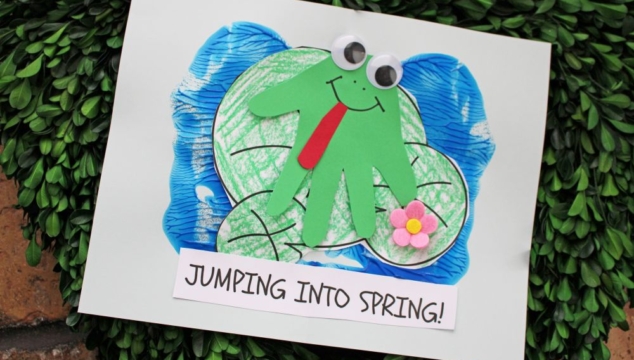 A handmade craft of a green frog with googly eyes and a red tongue, made from a handprint cutout, sits on a blue pond with lily pads. A small pink flower is attached, and a sign reads JUMPING INTO SPRING!.