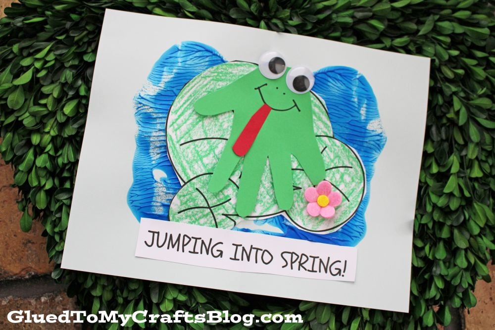 A handmade craft of a green frog with googly eyes and a red tongue, made from a handprint cutout, sits on a blue pond with lily pads. A small pink flower is attached, and a sign reads JUMPING INTO SPRING!.