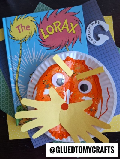 A paint splat Lorax craft project featuring a paper plate painted orange with googly eyes, a yellow paper mustache, and eyebrows, placed on top of The Lorax book. @GLUEDTOMYCRAFTS is labeled at the bottom.