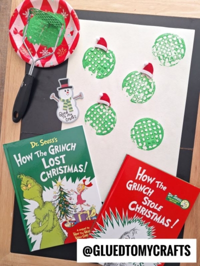 A craft setup inspired by Dr. Seuss’s Grinch features two Grinch-themed books, potato masher stamped craft green waffle prints with red Santa hats on white paper, a green spatula with a red plate, and a snowman cutout, all on a wooden table.