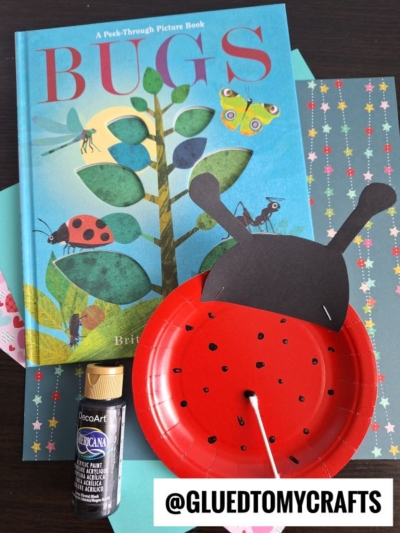 A childrens book titled Bugs, a ladybug craft made from a red paper plate with black spots and antennae, a bottle of black paint, and patterned paper sheets, all arranged on a table.