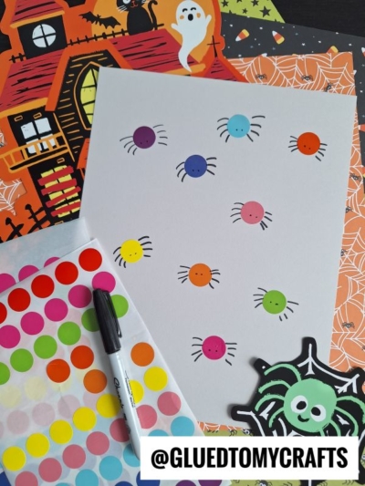A white sheet with colorful sticker dots turned into spiders using drawn legs—perfect for your INSECT CRAFT IDEAS. Surrounding the sheet are Halloween-themed papers, a black marker, dot stickers, and a cartoon spider cutout.