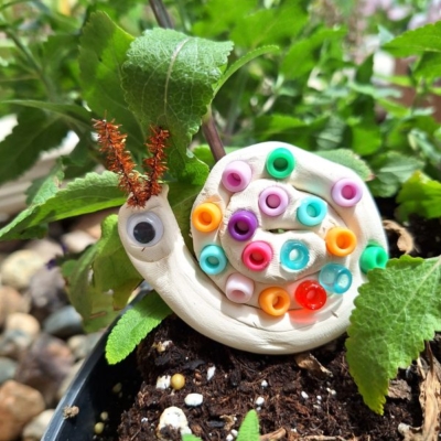 A snail craft made from white clay, colorful beads, a googly eye, and pipe cleaner antennae sits on soil among green leaves. Text at the bottom reads @GLUEDTOMYCRAFTS.