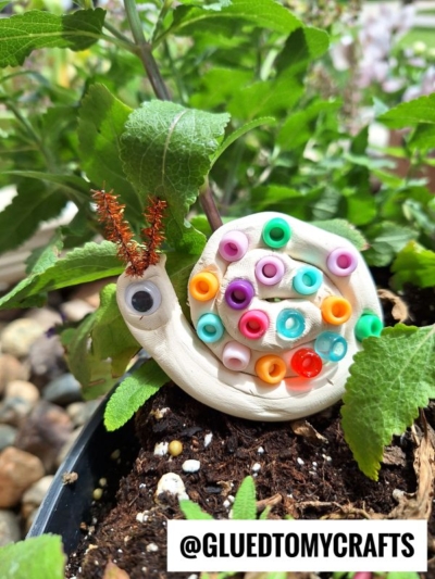 A snail craft made from white clay, colorful beads, a googly eye, and pipe cleaner antennae sits on soil among green leaves. Text at the bottom reads @GLUEDTOMYCRAFTS.