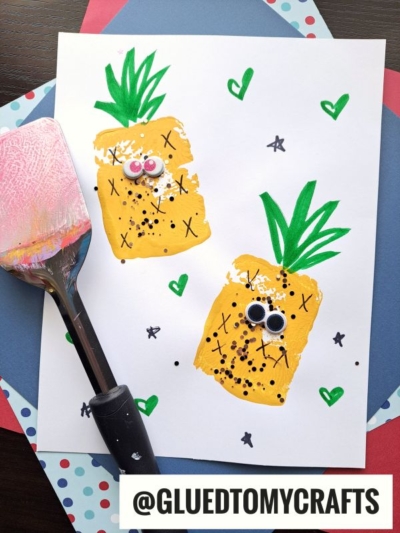 A painted craft showing two yellow pineapples with green tops, googly eyes, and hand-drawn details, on a white paper. A sponge brush with yellow paint lies beside it. Background includes polka-dotted and solid papers.