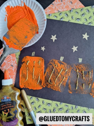 A fun FALL CRAFTS FOR KIDS scene with three orange painted pumpkins on black paper, a paint roller and plate, gold star stickers, glitter glue, a gold marker, plus patterned spiderweb and spider paper.