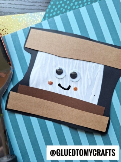 A spatula stamped smores craft features a googly-eyed, smiling paper s’more made from white, black, and brown paper, displayed on a blue striped background. The handle @GLUEDTOMYCRAFTS is at the bottom right.