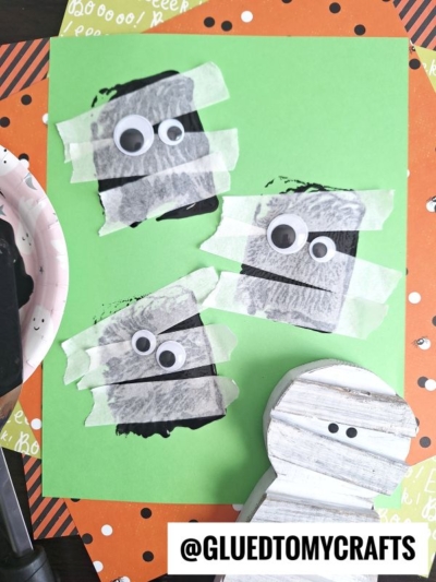 Three mummy faces made from black paint, white tape, and googly eyes—perfect for Sponge Painting Ideas for Kids—are arranged on green paper. A small wooden mummy figure sits nearby. The background is an orange polka-dot sheet. Text: @GLUEDTOMYCRAFTS.