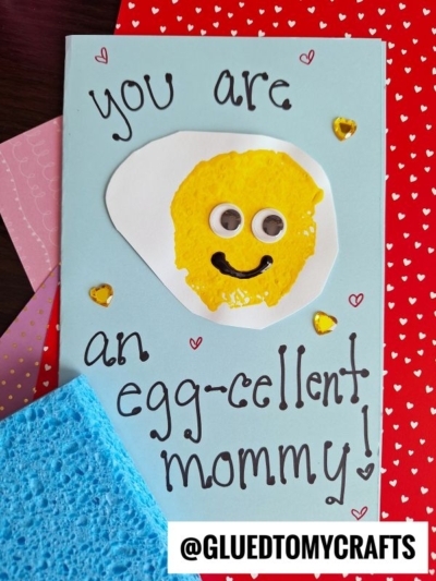 A handmade card with a painted fried egg featuring googly eyes and a smile, surrounded by hearts and gems. Perfect for Mother's Day or as inspiration for Sponge Painting Ideas for Kids! The text says, You are an egg-cellent mommy! @GLUEDTOMYCRAFTS appears at the bottom.