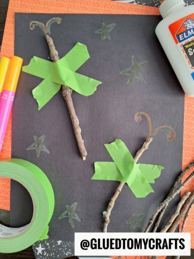 Two sticks taped to black paper with green tape to look like magic wands—perfect for FALL CRAFTS FOR KIDS. Hand-drawn stars shine in the background, with markers, glue, extra sticks, and a tape roll nearby for more crafting fun.