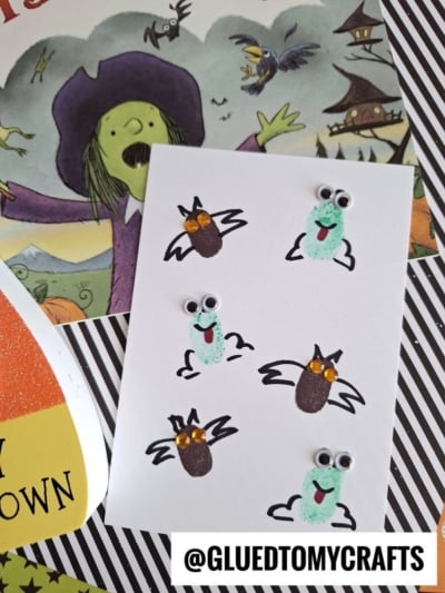 A handmade Halloween card showcases green and brown fingerprint creatures with googly eyes—some as bats, some as monsters—on white paper, surrounded by festive Halloween Crafts for Kids decorations and a whimsical witch illustration.
