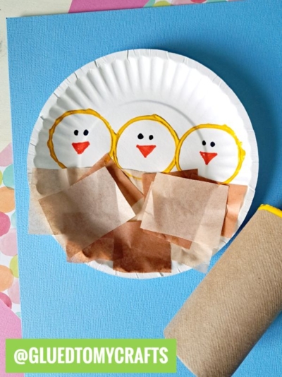 A paper plate decorated with three yellow chicks made from drawn circles and orange beaks, with brown paper squares as a nest, sits on blue paper alongside a cardboard tube. @GLUEDTOMYCRAFTS is written at the bottom.