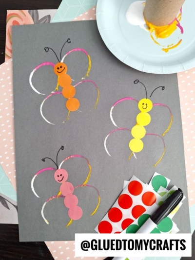 A craft project featuring three butterflies made from colored circle stickers on black paper, with wings painted in white, pink, and yellow. This fun coding label crafts idea also includes a marker and more stickers. Instagram: @GLUEDTOMYCRAFTS.