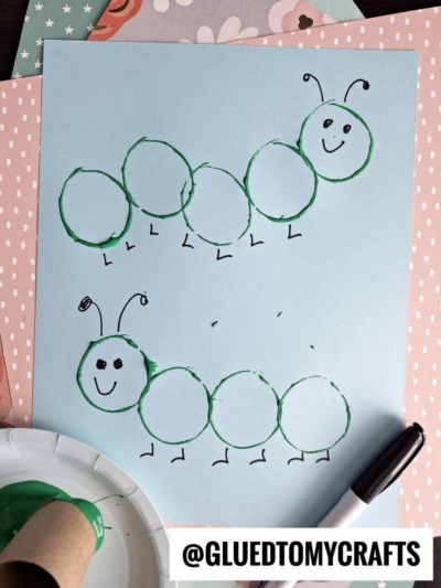 Two caterpillars made from green paint-stamped circles and drawn details are shown on blue paper—perfect for inspiring fun insect craft ideas, with a paint-covered cardboard tube, black marker, and patterned papers nearby.