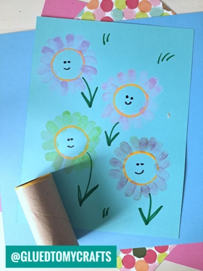 A blue sheet of paper with three colorful, smiling flower drawings made using paint and a cardboard tube stamp, placed among other craft papers. The handle @GLUEDTOMYCRAFTS appears at the bottom left.
