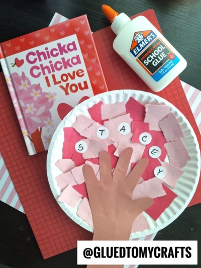 A children’s craft project features a paper plate with pink and red paper scraps, a cutout hand labeled STACEY, Elmer’s glue, the book Chicka Chicka I Love You, striped red paper—all perfect for cherry blossom tree crafts. @gluedtomycrafts.